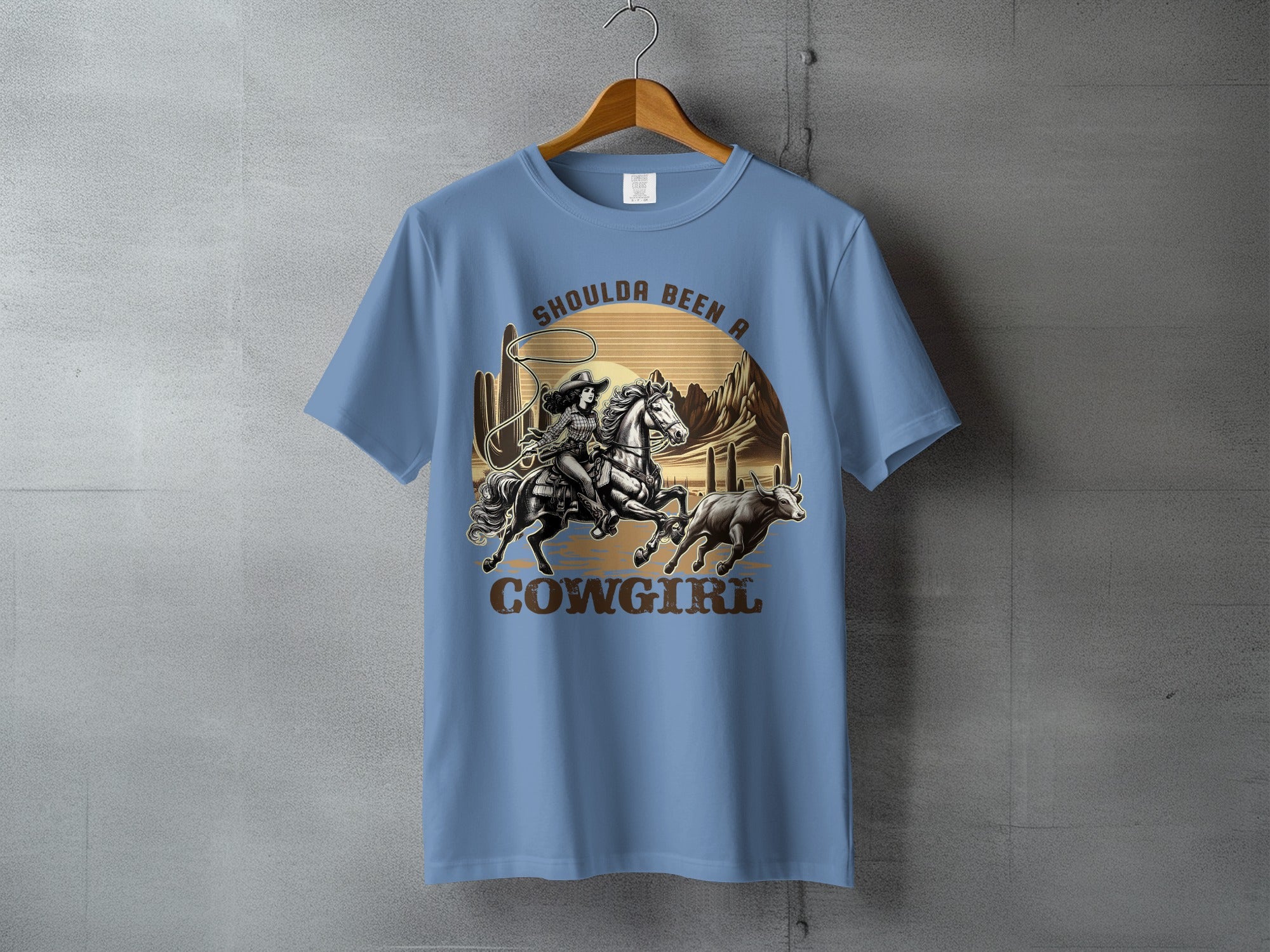 T-shirt with cowgirl on horseback lassoing a bull, desert scene, "Shoulda Been a Cowgirl" text.