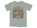 T-shirt with a rustic ranch wife design featuring a barn, boots, flowers, and bull skull.