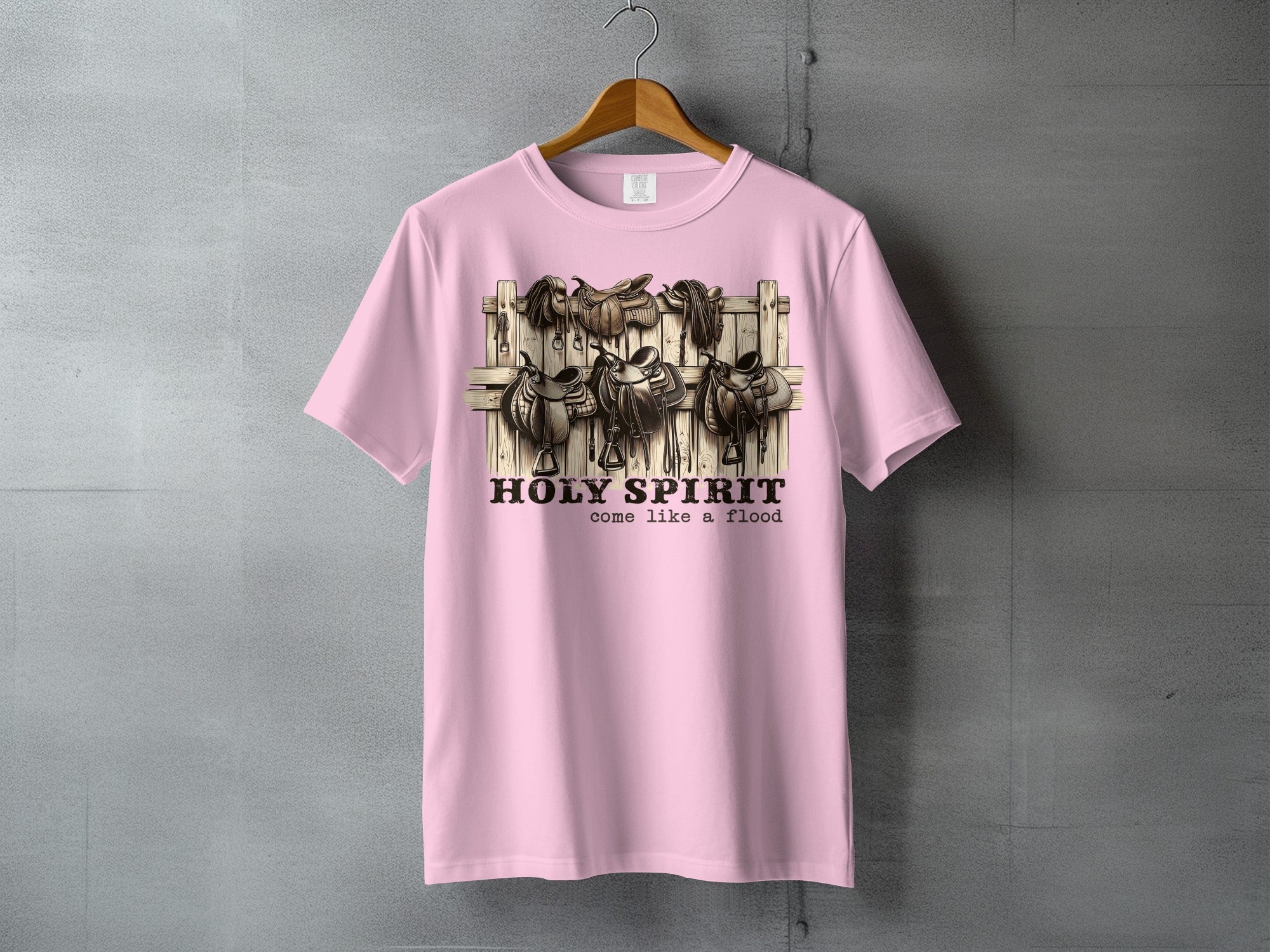 Pink t-shirt with western saddles hanging on a wooden fence, text reads "HOLY SPIRIT come like a flood."