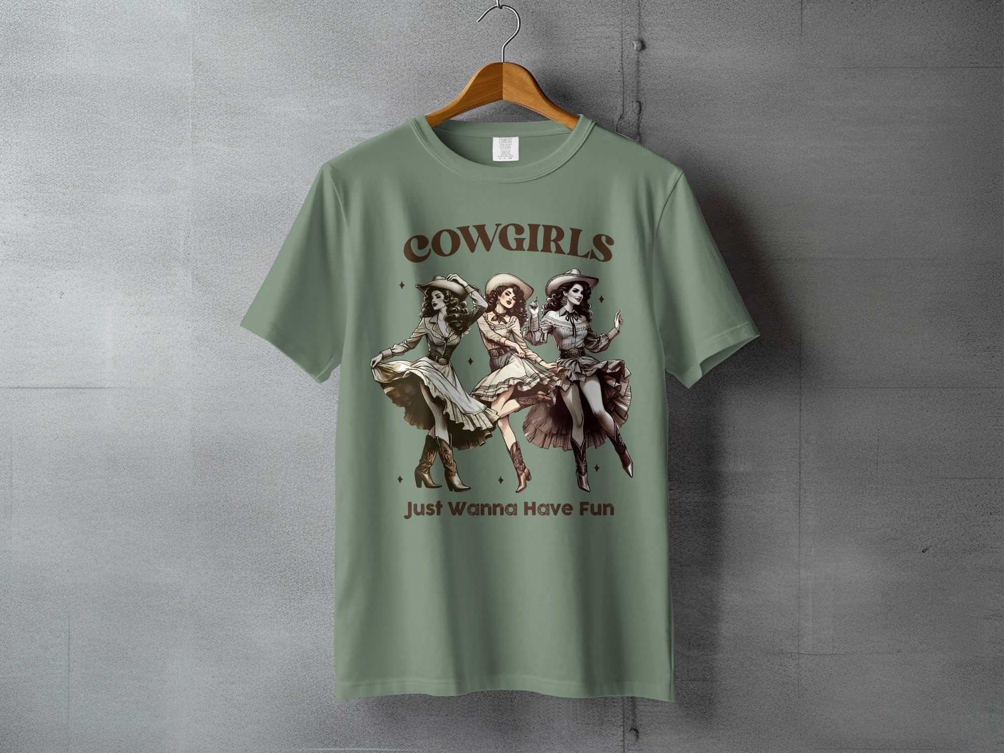 Three cowgirls dancing on a sage green t-shirt, reading "COWGIRLS Just Wanna Have Fun".